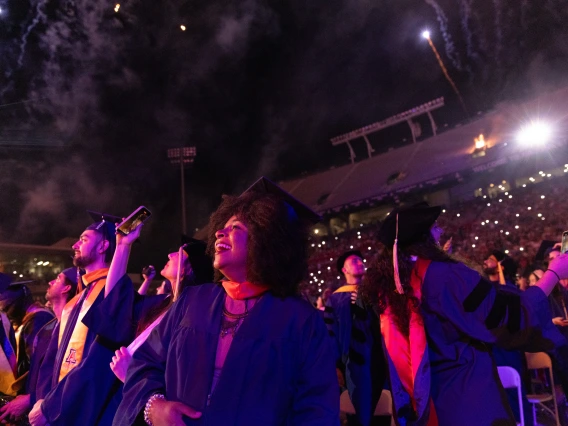 Graduates in academic robes look up at fireworks exploding above a stadium during a commencement ceremony.