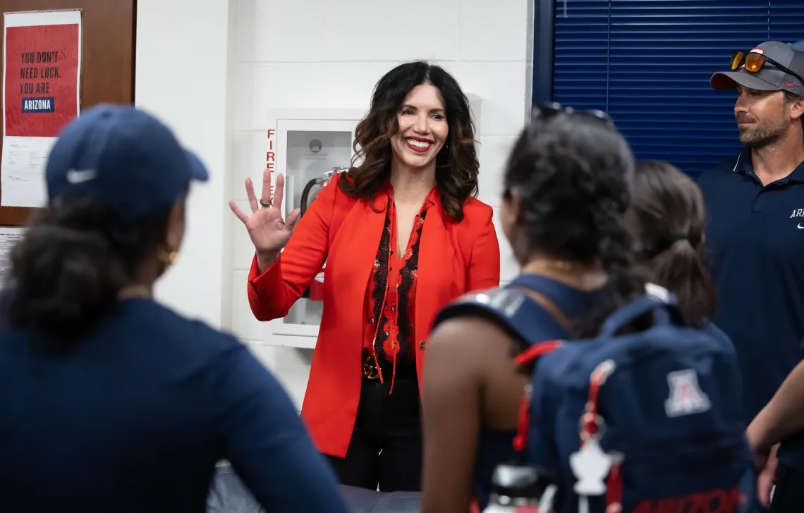 A photograph of a woman in a red blazer smiling and waving at student athletes