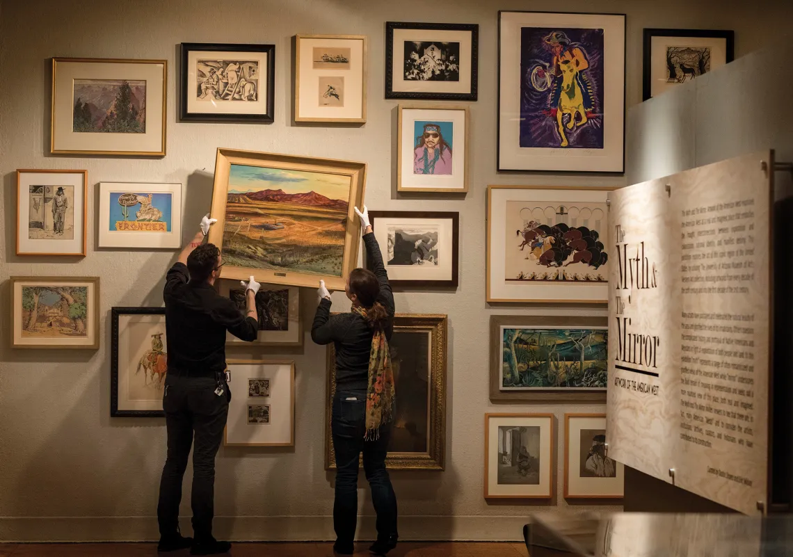 A photo of two individuals hanging up a painting on a wall surrounded by others