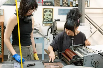 Image of professor Lisa Watanabe giving direction to student Scooter Curran on the Vandercook SP15.